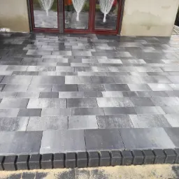 Ułożona, szara kostka brukowa przed wejściem do domu, obramowana czarnym, krawężnikiem. Częściowo widoczne okna z białymi firankami i elewacja budynku.