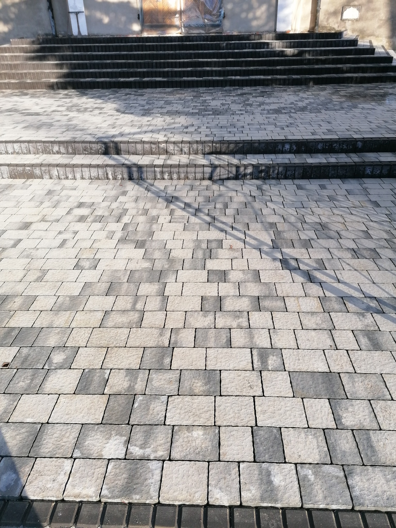 Schody wyłożone szarą kostką brukową, prowadzące do budynku z drewnianymi drzwiami zabezpieczonymi folią.