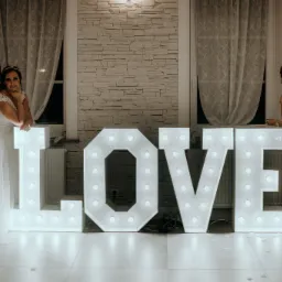 Panna młoda w białej sukni i druhna w bordowej sukni stoją po bokach podświetlanego napisu LOVE na tle kamiennej ściany i okien z białymi zasłonami.