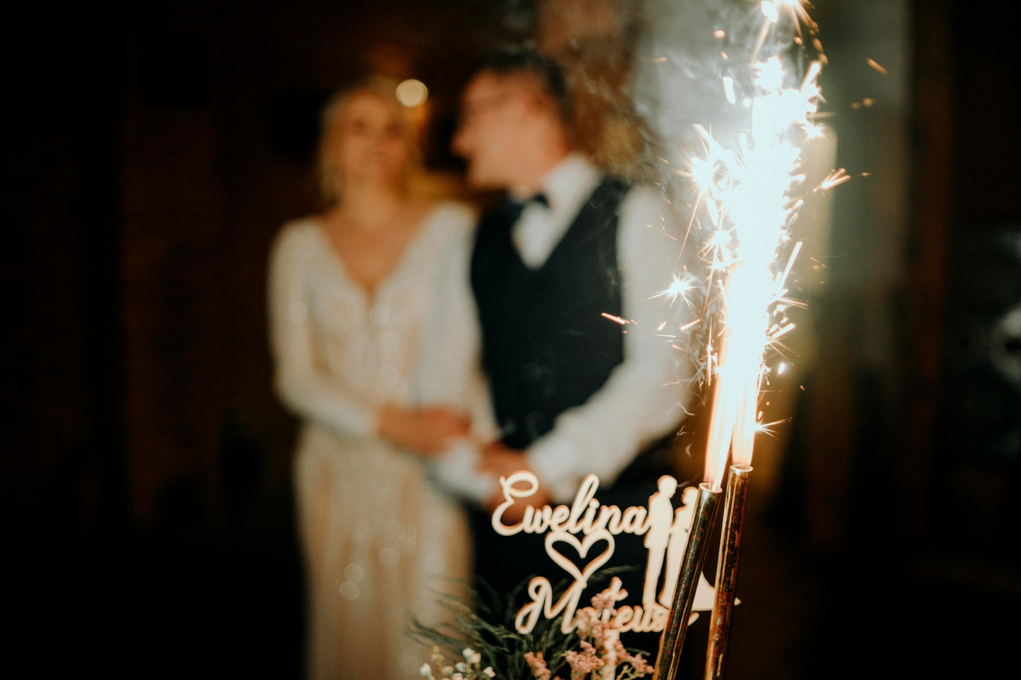 Młoda para w tle, przed nimi palące się zimne ognie i drewniany topper na tort z imionami 'Ewelina i Mateusz' oraz symbolem serca.