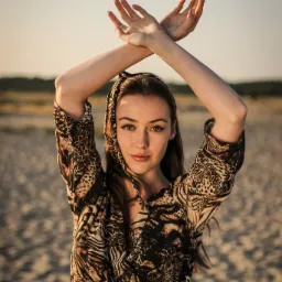 Portret młodej kobiety w sukience w zwierzęcy wzór i chuście na głowie, dłonie skrzyżowane nad głową na tle piaszczystej plaży i jasnego nieba.