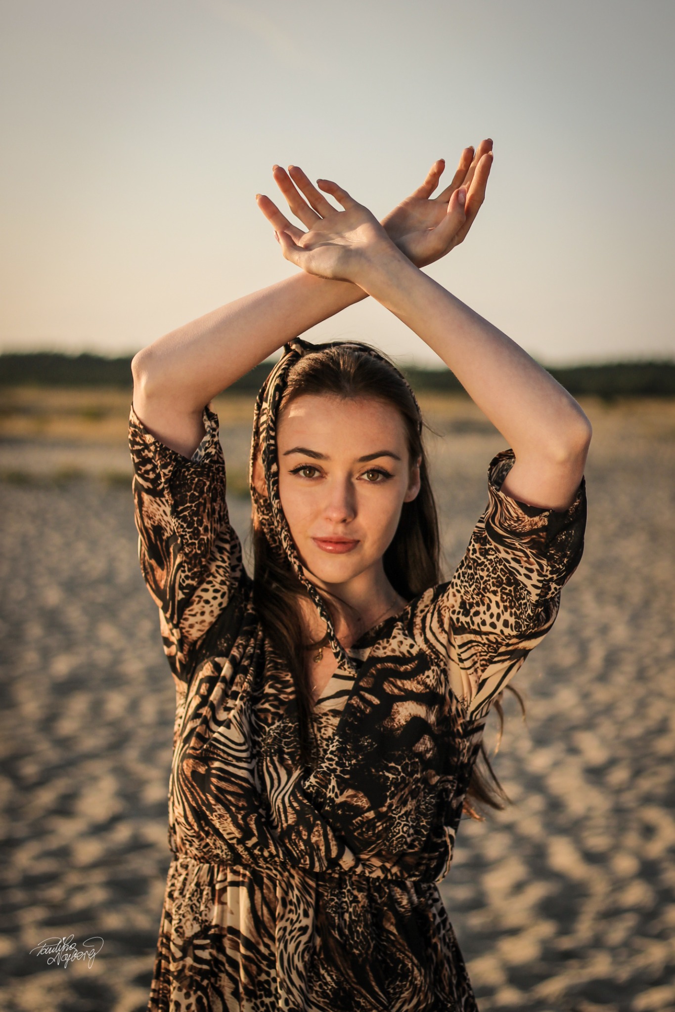 Portret młodej kobiety w sukience w zwierzęcy wzór i chuście na głowie, dłonie skrzyżowane nad głową na tle piaszczystej plaży i jasnego nieba.