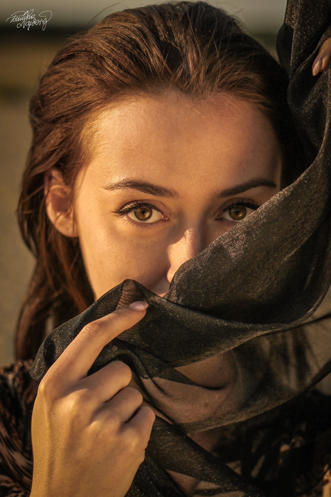 Portret kobiety z brązowymi włosami, zmysłowo spoglądającej znad czarnej, połyskującej tkaniny, która częściowo zasłania jej twarz.