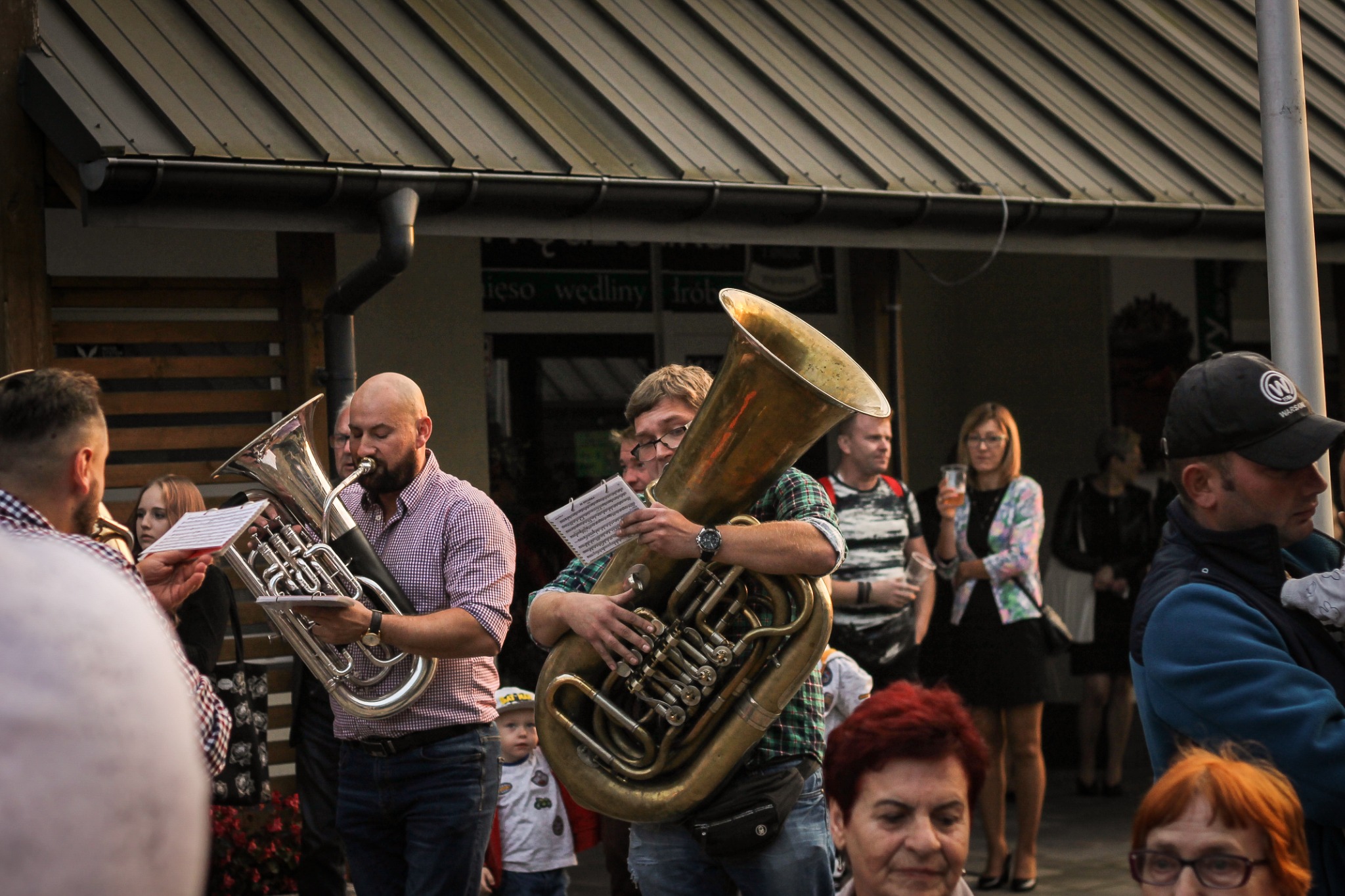 Zespół muzyczny grający na instrumentach dętych, w tle widzoczni ludzie i budynek.