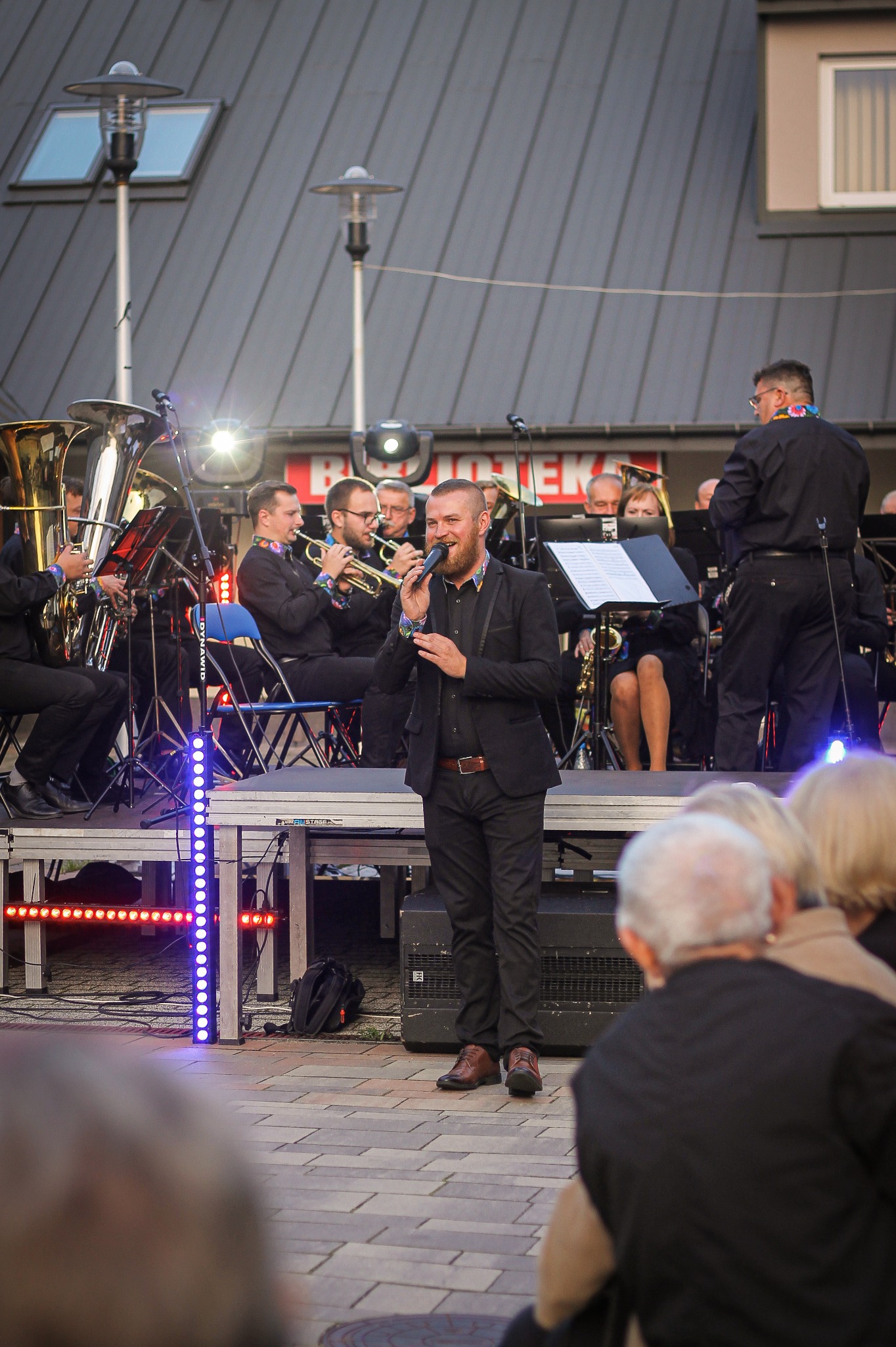 Wokalista z brodą w czarnym garniturze śpiewa do mikrofonu na scenie podczas koncertu plenerowego z orkiestrą dętą w tle, widownia obserwuje występ.