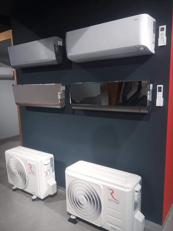 Ekspozycja klimatyzatorów różnych kolorów (biały, szary, czarny) i modeli na ciemnej ścianie, z jednostkami zewnętrznymi na podłodze.