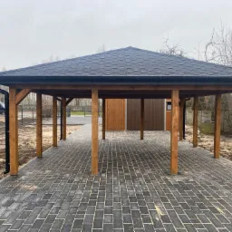 Zadaszona wiata drewniana z czarnym dachem bitumicznym na posesji z kostką brukową, widok na dom i ogrodzenie.