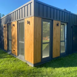 Nowoczesny, parterowy dom modułowy z drewnianą elewacją i grafitowym dachem z blachy trapezowej, oświetlony kinkietami, otoczony trawnikiem w słoneczny dzień.