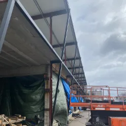 Wznoszenie konstrukcji budynku z betonu i stali, widoczna podnośnik nożycowy JLG, zabezpieczenia z plandek i sterta drewna na placu budowy.