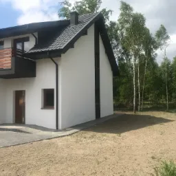 Nowoczesny dom jednorodzinny z białymi ścianami, ciemną dachówką i drewnianym balkonem, otoczony drzewami i terenem przygotowanym pod trawnik.