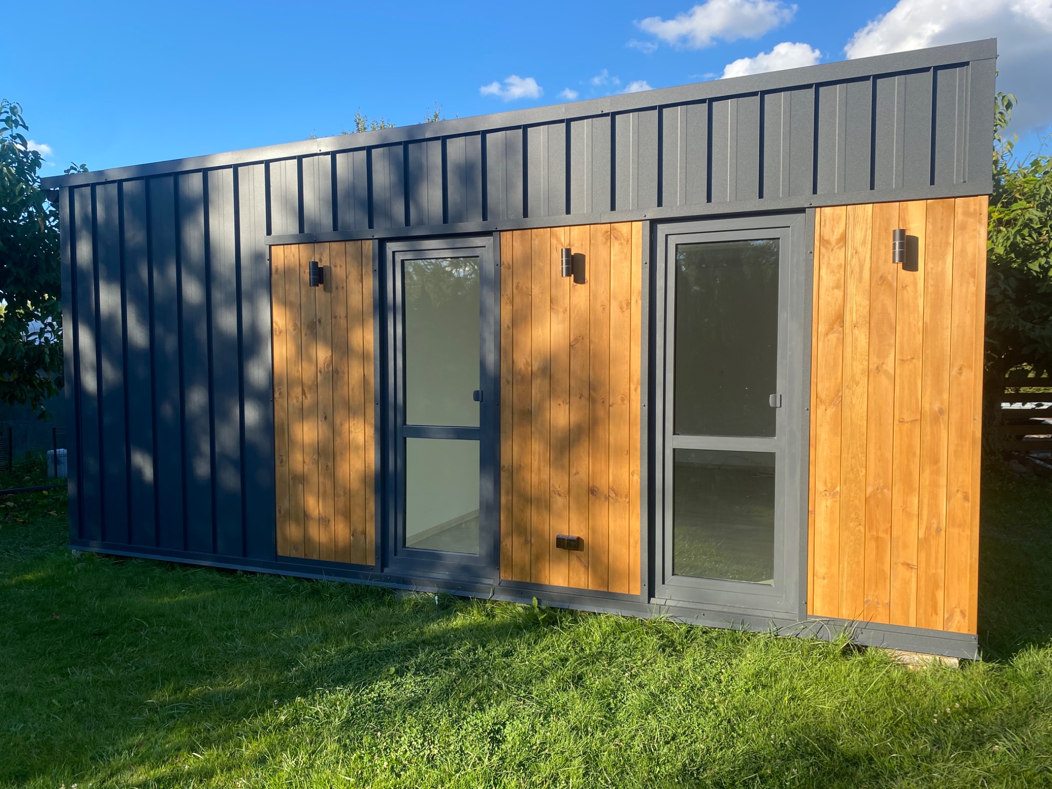Nowoczesny, szary budynek modułowy z drewnianymi panelami i oświetleniem zewnętrznym, otoczony zielonym trawnikiem w słoneczny dzień.