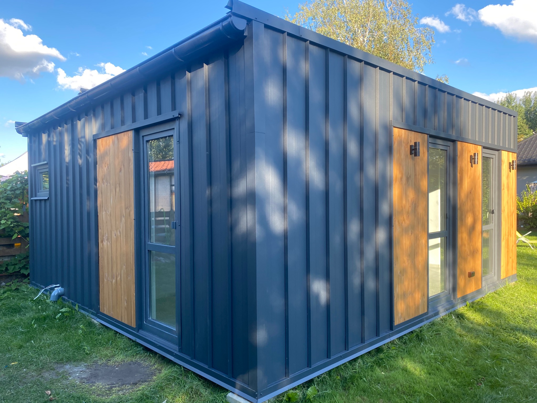 Nowoczesny, szary dom modułowy z drewnianymi akcentami, widoczny z zewnątrz na tle trawnika i drzew.