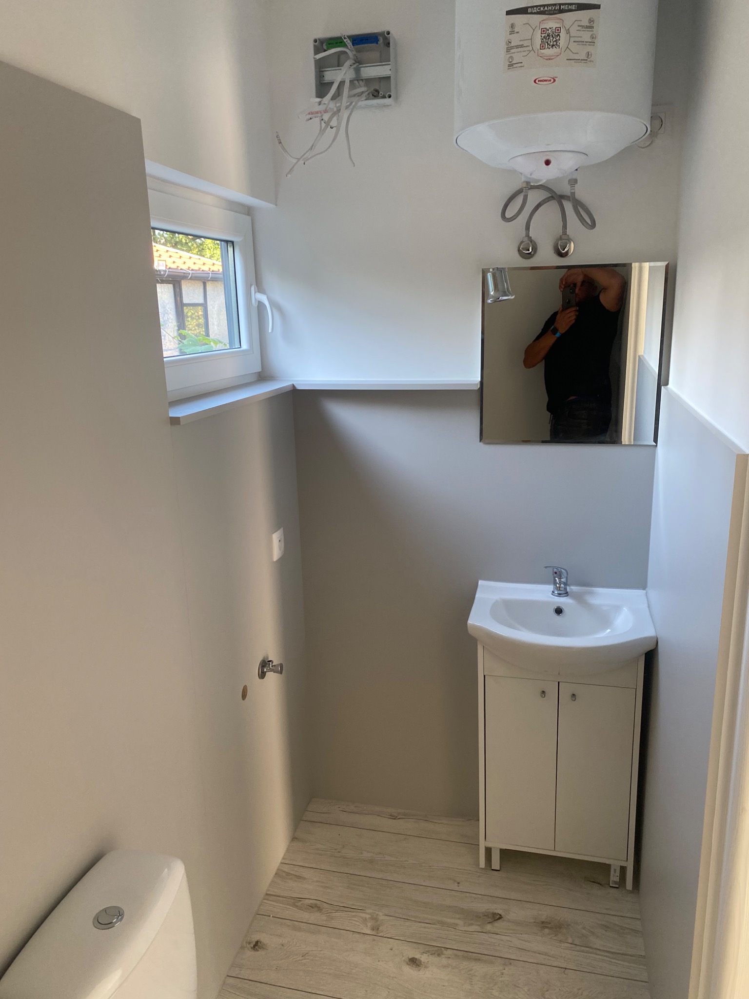 Wnętrze łazienki w trakcie wykańczania: biała umywalka z szafką, toaleta, bojler elektryczny z podłączonymi rurami, lustro z odbiciem fotografa, okno z widokiem na zewnątrz, widoczne przewody...