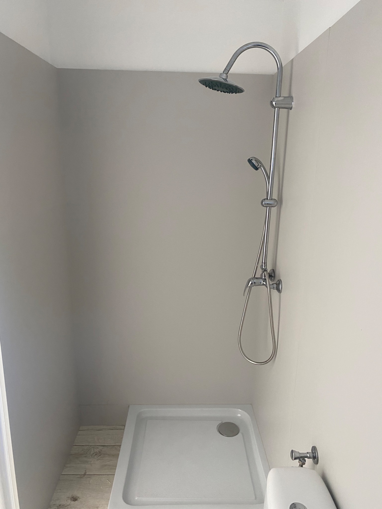 Kabina prysznicowa z deszczownicą i słuchawką, zamontowana w narożniku łazienki z szarymi ścianami i brodzikiem. Widoczny fragment toalety z zaworem.