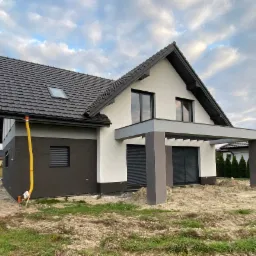 Nowoczesny dom jednorodzinny z dwukolorową elewacją (biały tynk i ciemny brąz), ciemną dachówką i szarą pergolą, widoczny żółty przewód wentylacyjny na elewacji, teren wokół domu jeszcze...