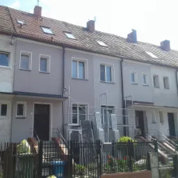 Szeregówka domów po częściowym malowaniu elewacji na szaro, z widocznymi rusztowaniami i elementami przygotowanymi do dalszych prac.