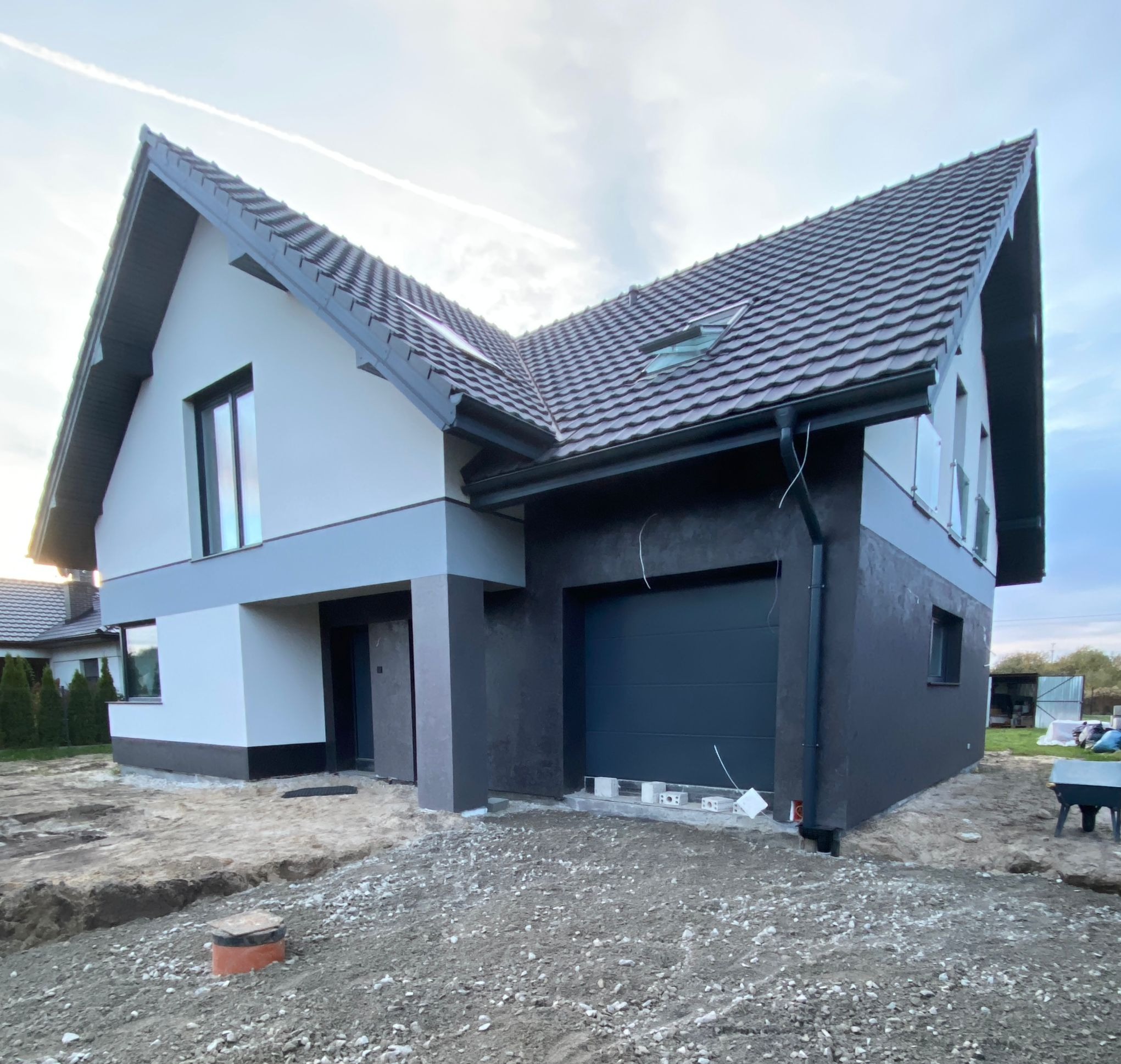 Dwukondygnacyjny dom jednorodzinny z szarą elewacją i ciemną stolarką okienną, widok z zewnątrz na częściowo zagospodarowany teren przed budynkiem.