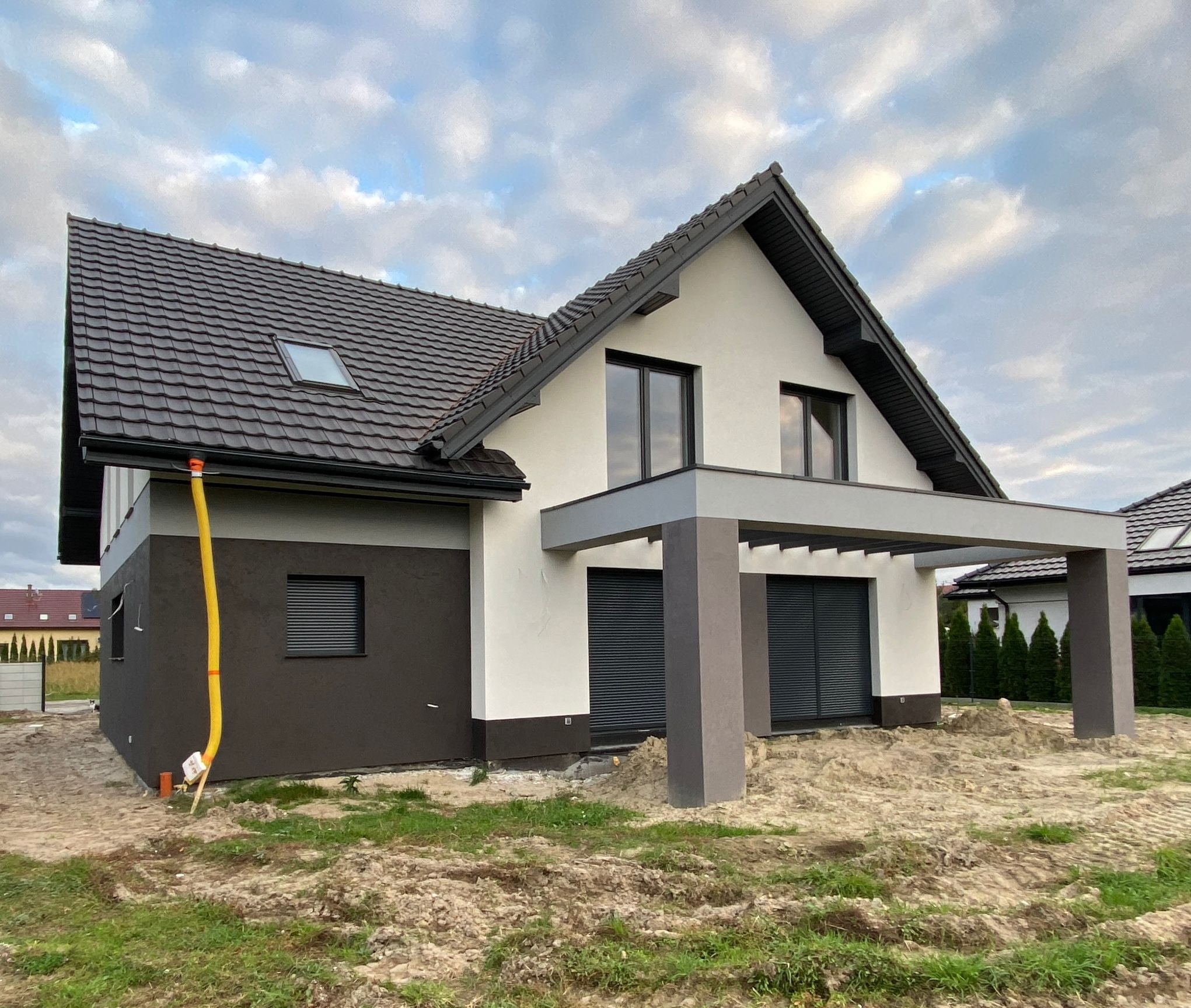 Nowoczesny dom jednorodzinny z dwukolorową elewacją (biały tynk i ciemny brąz), ciemną dachówką i szarą pergolą, widoczny żółty przewód wentylacyjny na elewacji, teren wokół domu jeszcze...