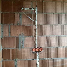 Mur z cegły z bruzdami wypełnionymi pianką montażową, pomarańczowe puszki instalacyjne w ścianie, kabel wystający z bruzdy.
