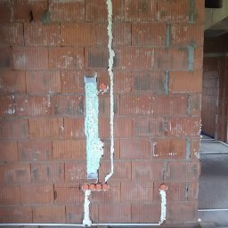 Usługi elektryczne Dawid Mieseler - Murowana ściana z bruzdami wypełnionymi pianką montażową, w których umieszczono przewody elektryczne i pomarańczowe puszki instalacyjne. Widoczne fragmenty podłogi i surowego stanu budynku.