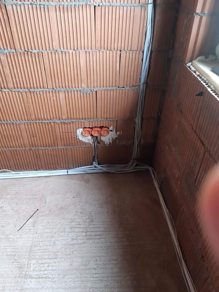 Surowy stan instalacji elektrycznej w nowym budynku: kable prowadzone wzdłuż ściany z cegły, widoczne puszki elektryczne w ścianie, podłoga betonowa w trakcie prac.