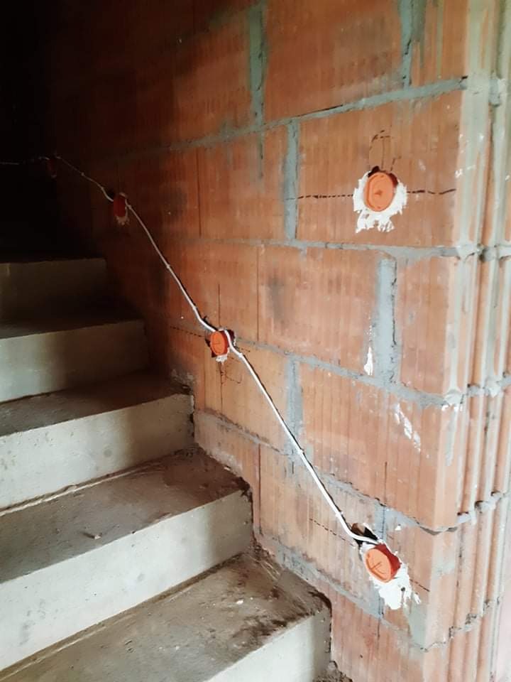 Instalacja elektryczna w surowym stanie: przewód prowadzony po ścianie z cegły przy schodach, widoczne pomarańczowe puszki instalacyjne.