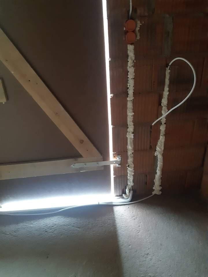 Montaż instalacji elektrycznej w surowym wnętrzu: przewody w pianie montażowej przy ceglanej ścianie, puszki elektryczne i drewniana konstrukcja pod sufitem.