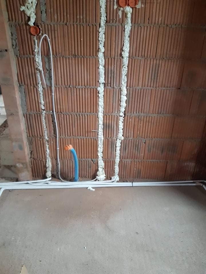 Surowa ściana z cegły dziurawki z instalacją elektryczną w białych peszlach, zabezpieczona pianką montażową, widoczne puszki instalacyjne w kolorze pomarańczowym i fragment niebieskiej rury...