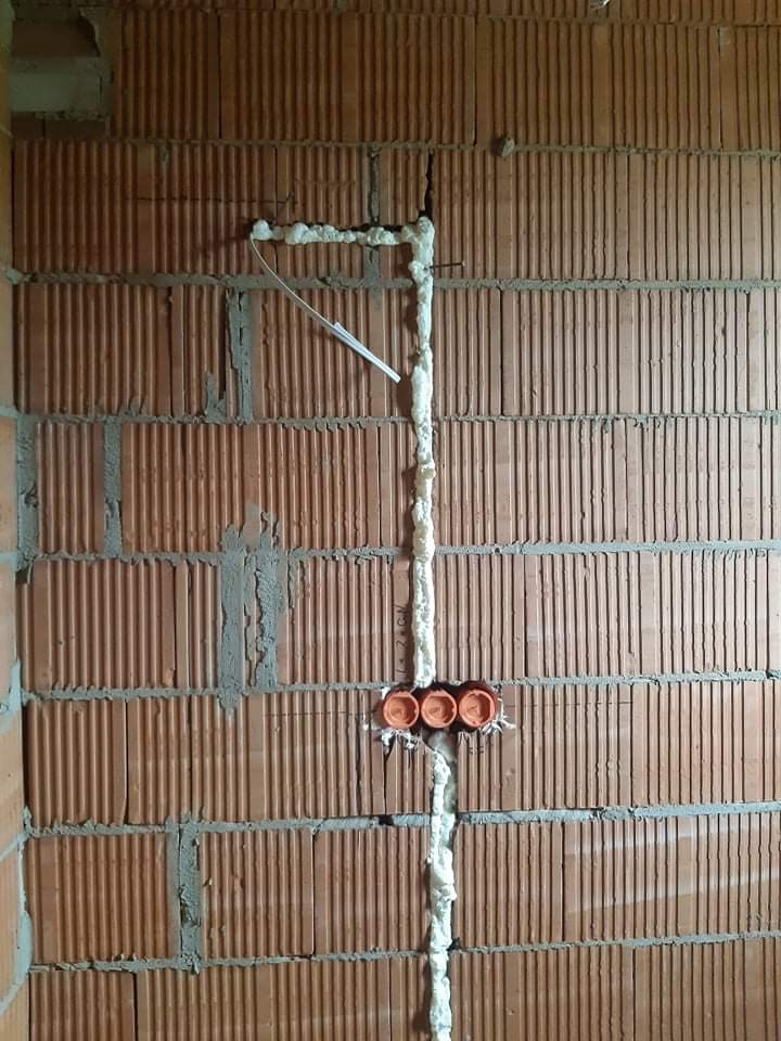 Mur z cegły z bruzdami wypełnionymi pianką montażową, pomarańczowe puszki instalacyjne w ścianie, kabel wystający z bruzdy.