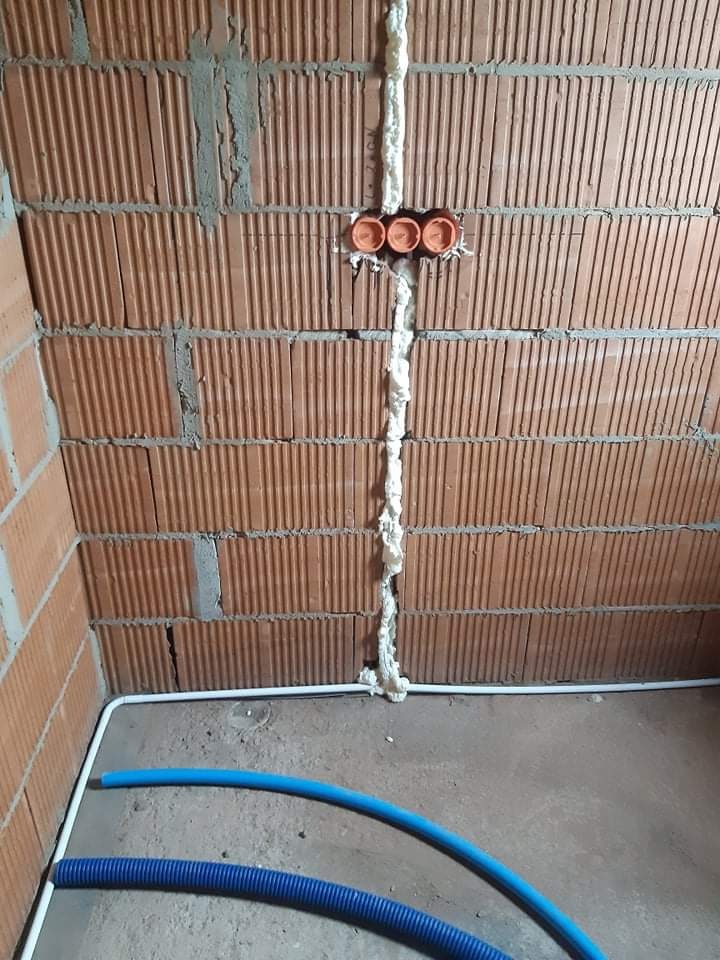 Murowana ściana z cegły dziurawki z trzema pomarańczowymi puszkami instalacyjnymi, zabezpieczonymi pianką montażową, oraz niebieskie rury karbowane na podłodze.