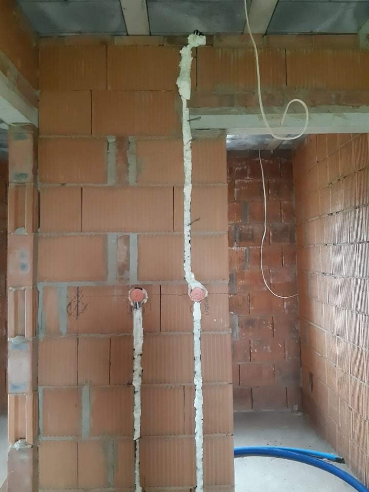 Instalacja elektryczna w bruzdach wypełnionych pianką montażową w surowym murze z czerwonej cegły dziurawki, widoczne wystające przewody i puszki instalacyjne.