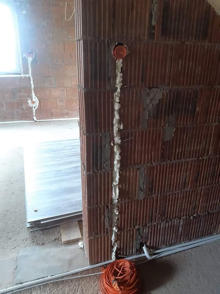 Montaż puszki elektrycznej w ścianie z cegły, zabezpieczonej pianką montażową, widoczne przewody i zwój pomarańczowego kabla.