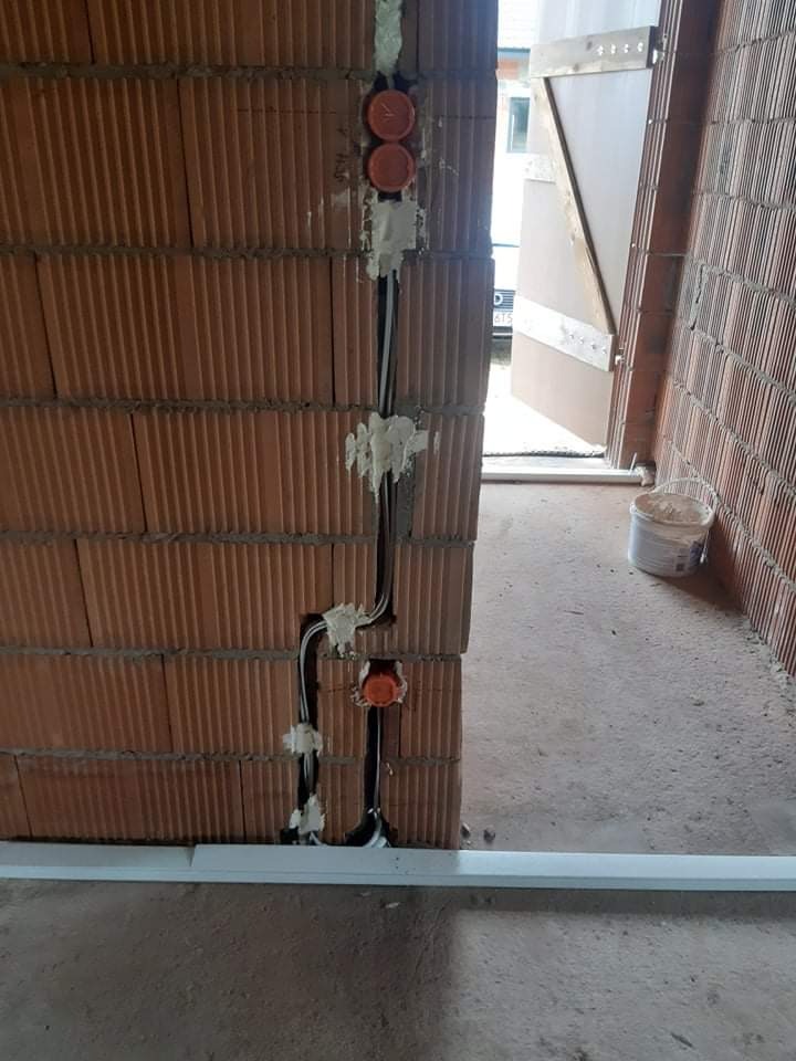 Wewnętrzna ściana z cegły z pomarańczowymi puszkami instalacyjnymi i przewodami zabezpieczonymi pianką montażową, widok na otwarte drzwi i fragment podłogi z listwą przypodłogową.