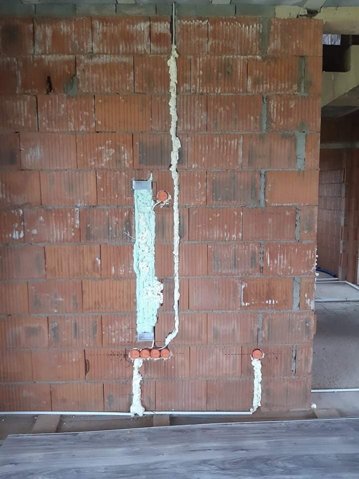 Murowana ściana z bruzdami wypełnionymi pianką montażową, w których umieszczono przewody elektryczne i pomarańczowe puszki instalacyjne. Widoczne fragmenty podłogi i surowego stanu budynku.
