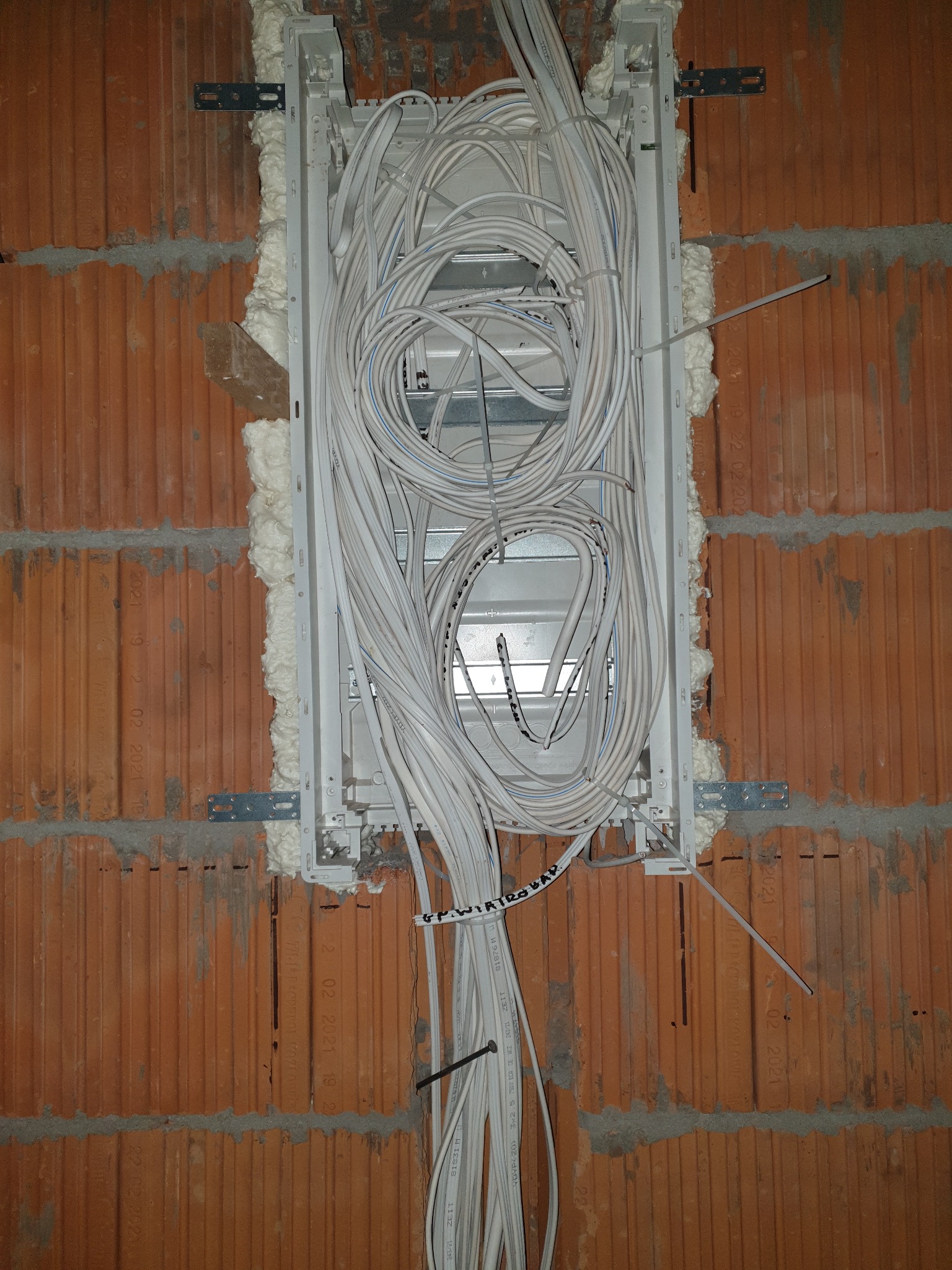 Wnęka w surowej ścianie z cegły wypełniona białą pianką montażową, wewnątrz której umieszczono metalową skrzynkę z licznymi białymi kablami elektrycznymi spiętymi opaską. Widoczne mocowania...