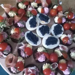 Różnorodne przekąski na drewnianej tacy: jajka z czarnym kawiorem, mini tartaletki z kremem, szynką parmeńską i pomidorkami koktajlowymi, udekorowane koperkiem.