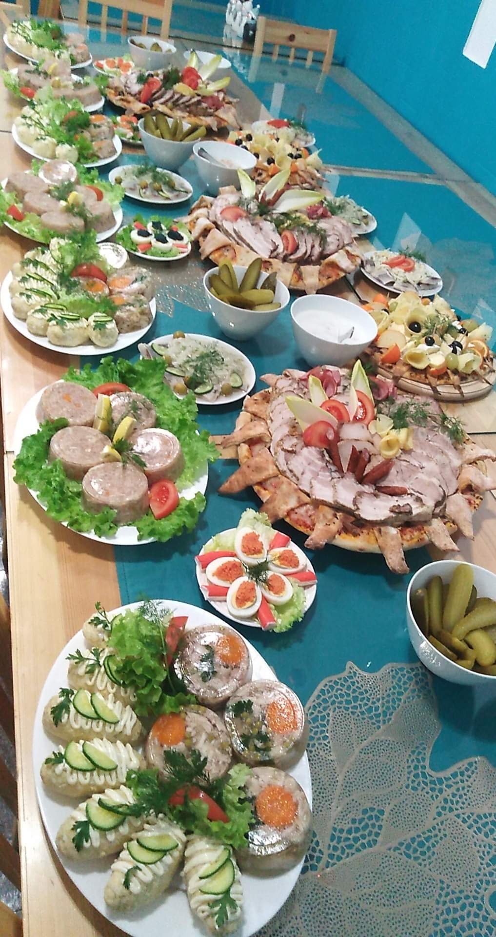 Stół zastawiony różnorodnymi daniami: galaretki mięsne z warzywami, jajka faszerowane, wędliny na pizzy, sałatki i ogórki konserwowe w miseczkach, gotowe do serwowania.