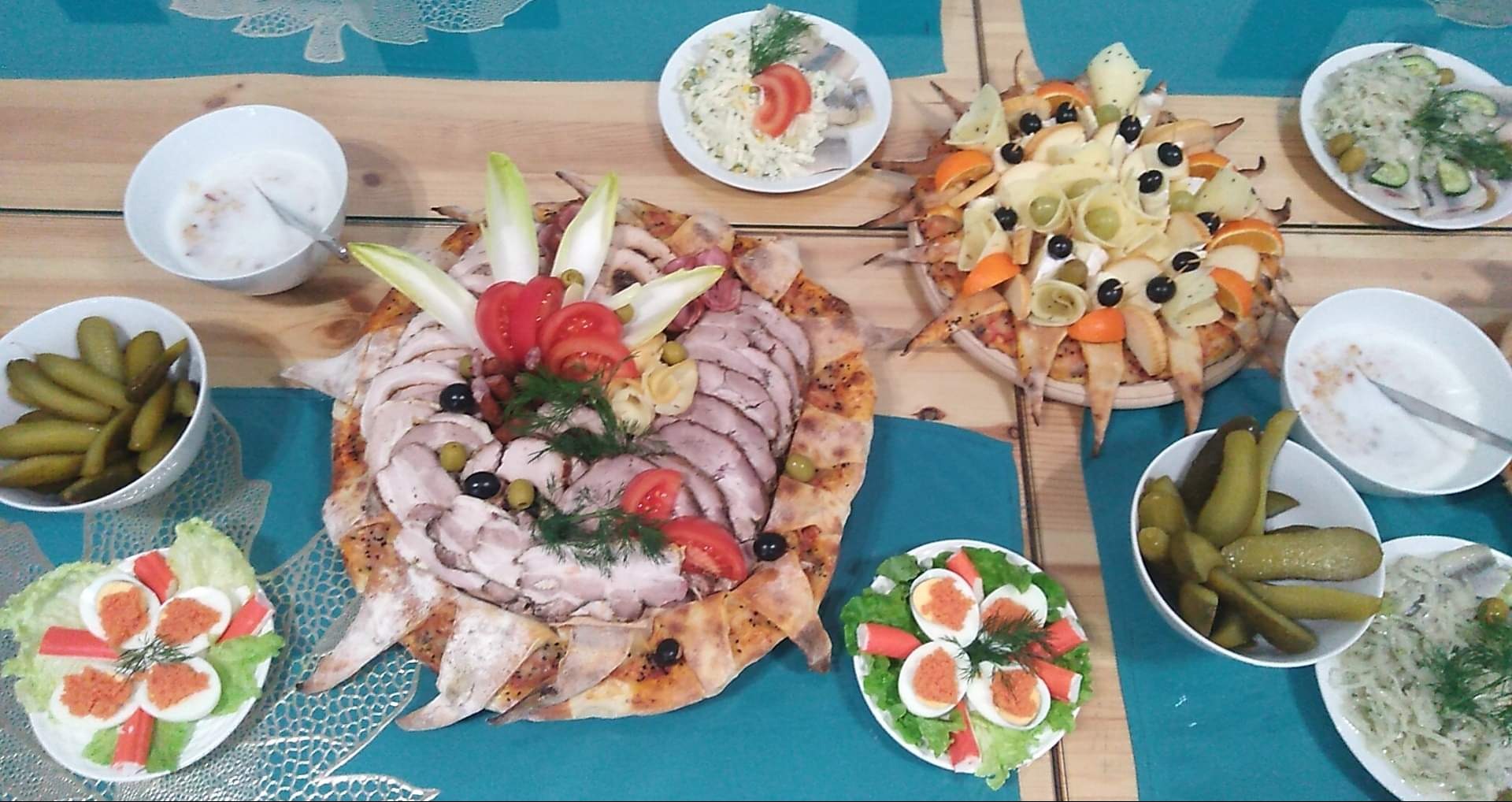 Uroczysty stół zastawiony różnorodnymi potrawami: patery z wędlinami, serem i owocami ułożone na pizzy, sałatki, ogórki kiszone, sos w miseczkach, dekoracje z warzyw i ziół.