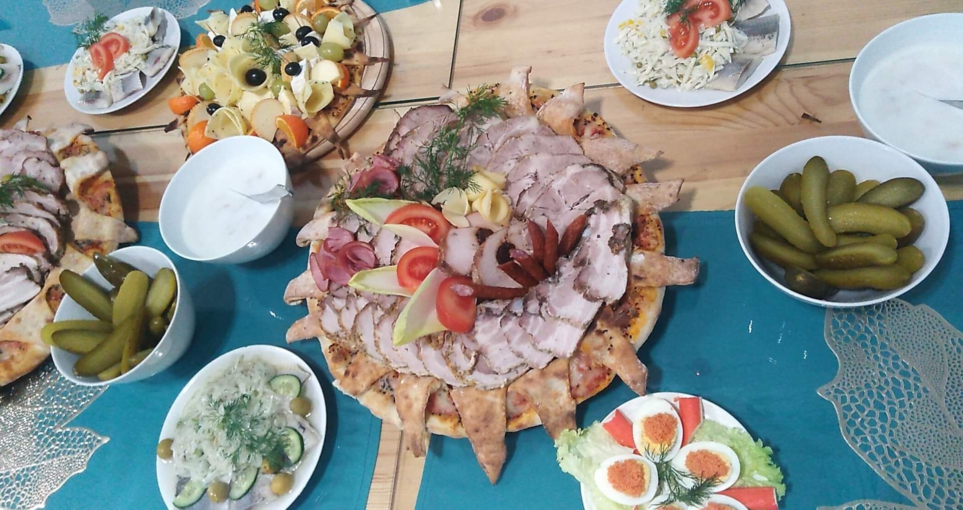 Bogato zastawiony stół z różnorodnymi przekąskami: talerz wędlin na cieście, sałatki, śledzie, ogórki kiszone, jajka faszerowane, serwowane na drewnianym stole z turkusową serwetą z ażurowym wzorem.