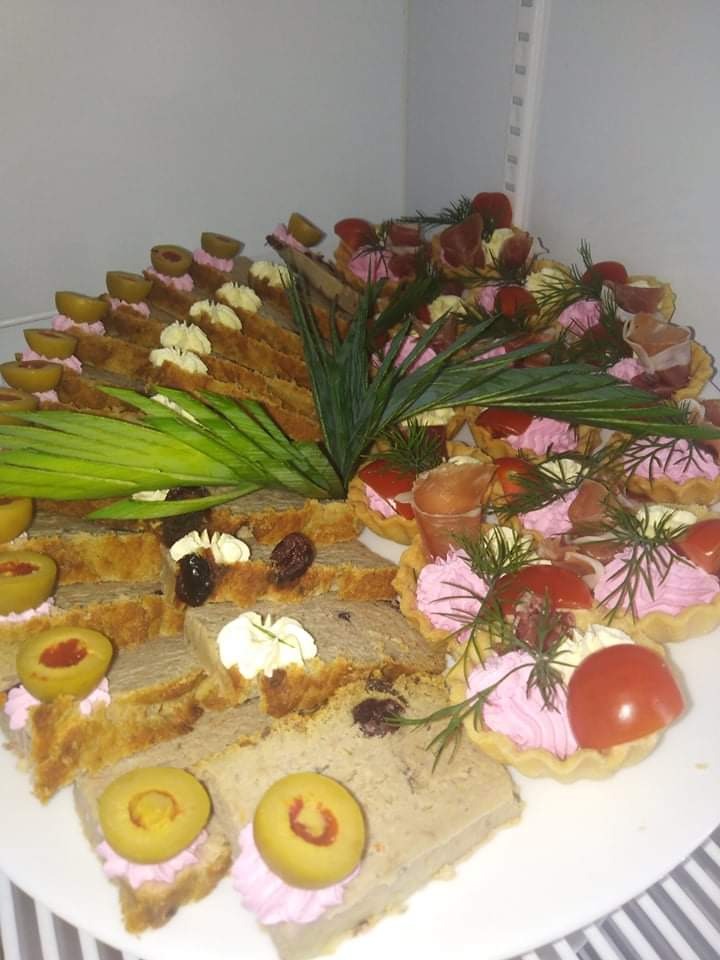 Dekoracyjnie ułożone przekąski cateringowe: pasztet z oliwkami, tartaletki z kremem i pomidorem, wędlina w rożkach, całość udekorowana zielonymi gałązkami.