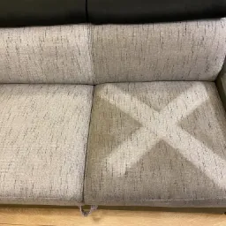 Częściowo wyczyszczona szara kanapa z widocznym znakiem w kształcie litery X, wskazującym obszar po praniu.