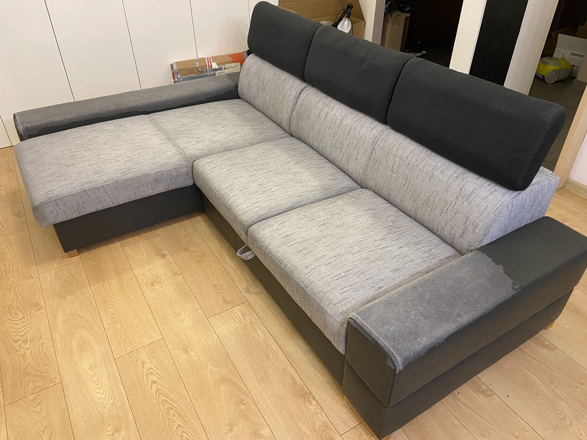 Szara, narożna kanapa z czarnymi zagłówkami na drewnianej podłodze. Widoczny fragment białej szafy i kartonów w tle.