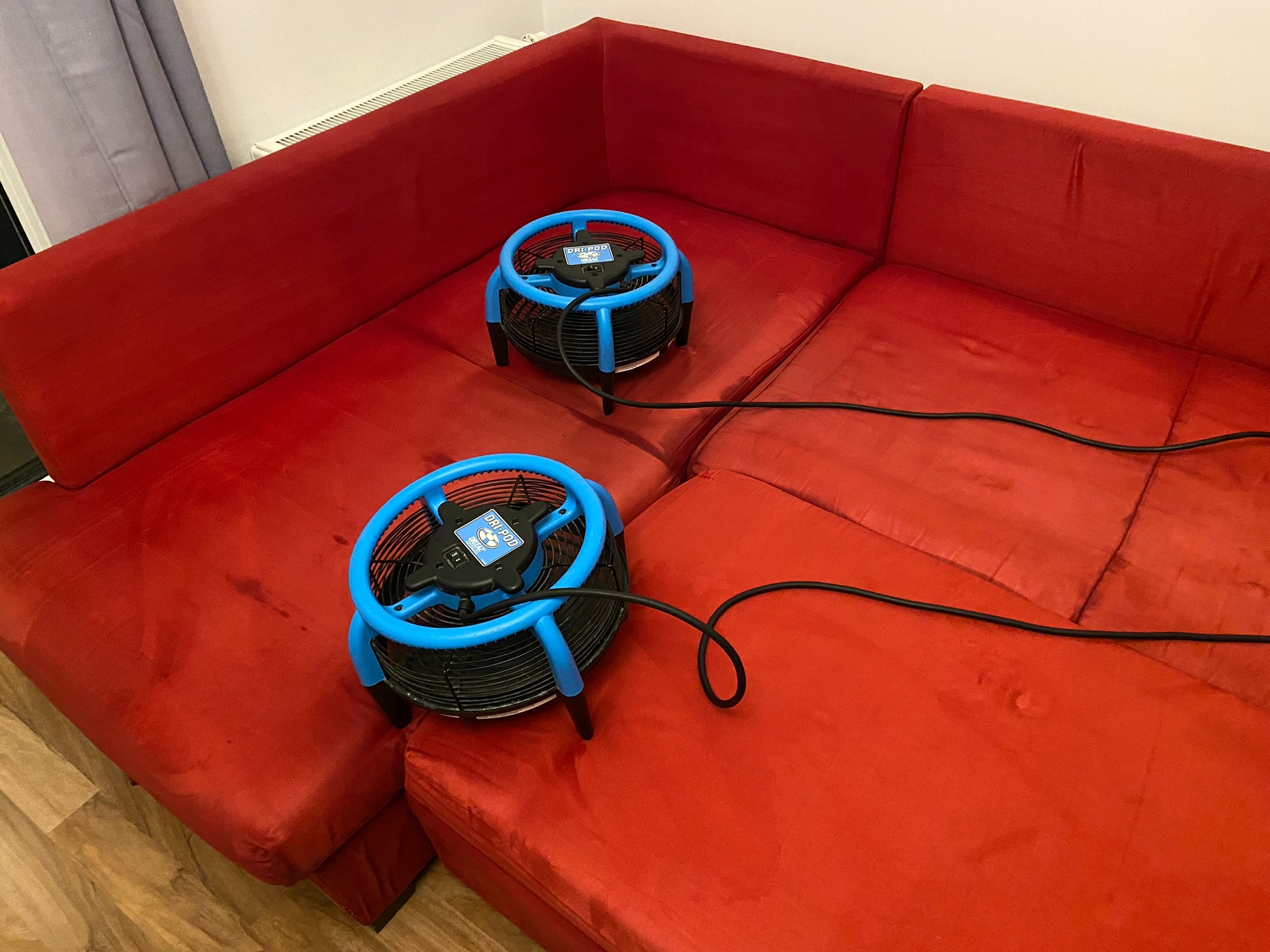 Dwa niebieskie wentylatory osuszające, marki Dri-Pod, stoją na czerwonej, modułowej kanapie. Widoczne ślady po czyszczeniu na materiale.
