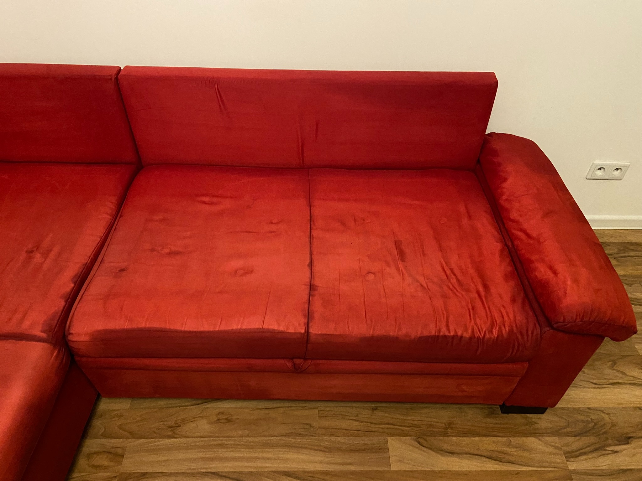 Czerwona, narożna kanapa z widocznymi śladami użytkowania na siedzisku, stojąca na panelach podłogowych w pobliżu gniazdka elektrycznego.