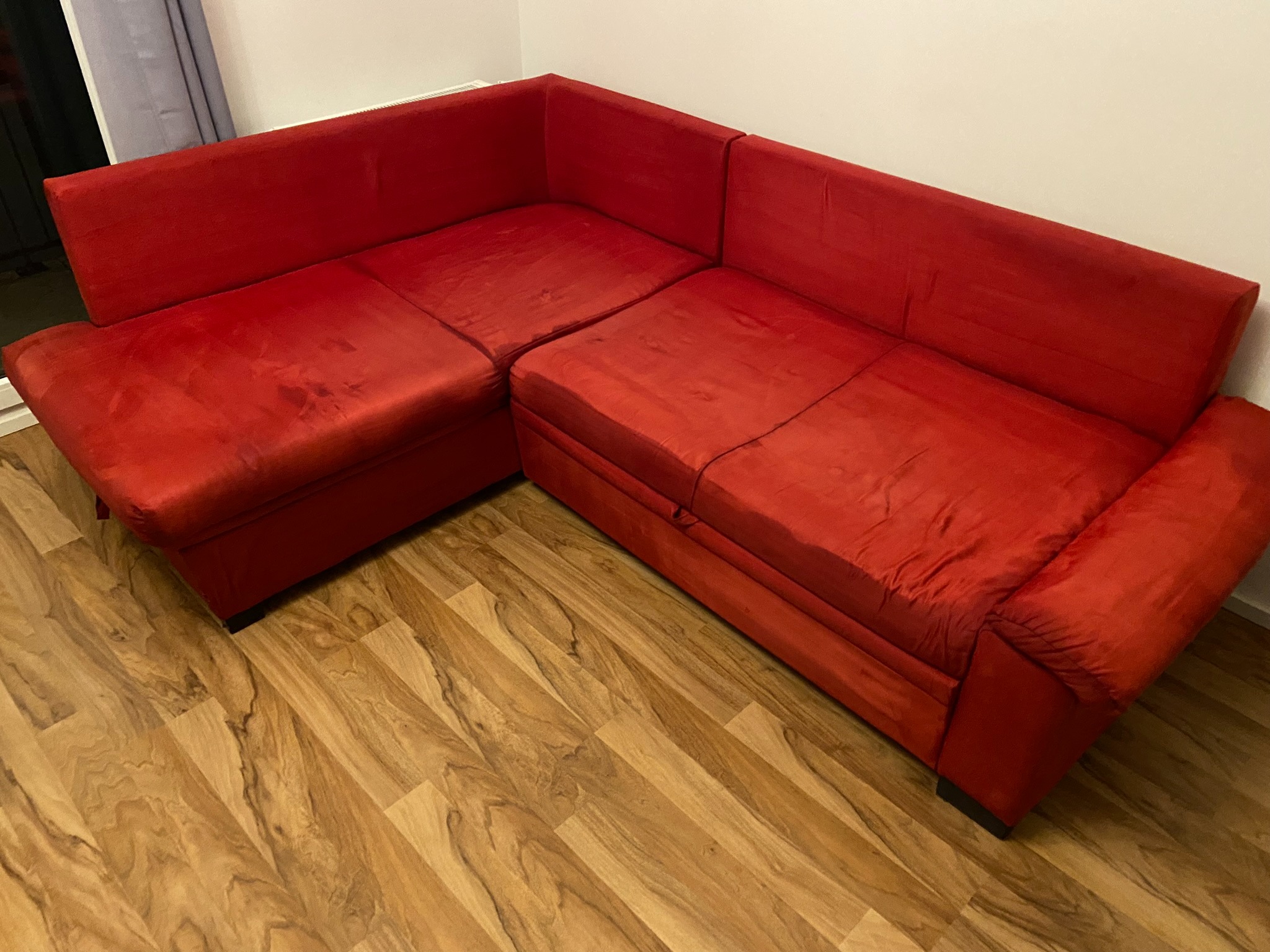 Czerwona, narożna kanapa z materiału o widocznych śladach użytkowania, stojąca na podłodze z paneli imitujących drewno.