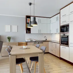 Jasne wnętrze salonu z aneksem kuchennym w Mysłowicach. Widoczny st&oacute;ł z krzesłami, fotel uszak, szafki oraz zabudowa kuchenna z białymi frontami i drewnianymi akcentami. Nad stołem wiszą dwie szare...