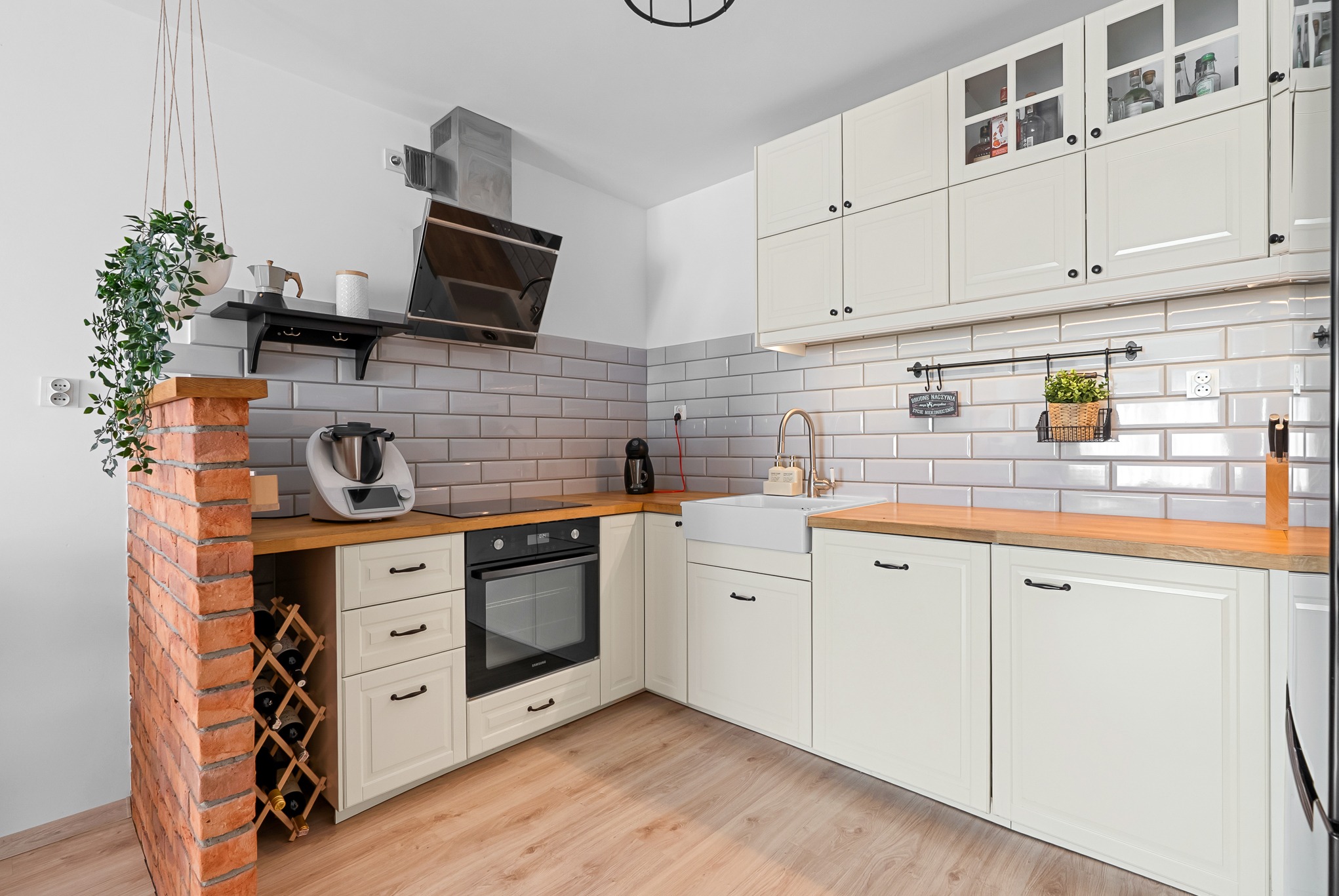 Jasna kuchnia w stylu rustykalnym z ceglanym elementem, drewnianym blatem i białymi szafkami. Widoczny ekspres do kawy i stojak na wino.