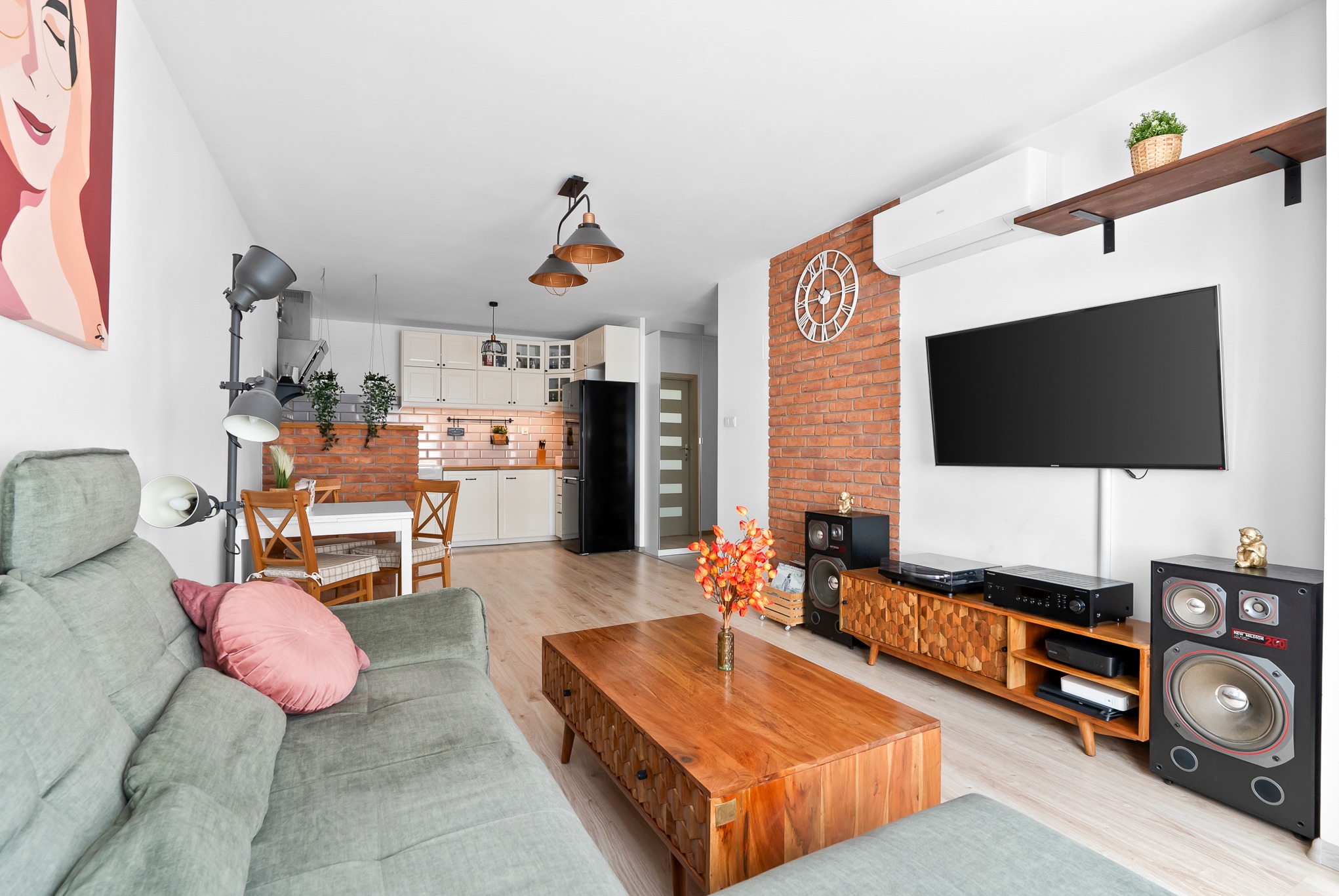 Jasne wnętrze: salon z aneksem kuchennym. Wygodna sofa, drewniany stolik kawowy, ceglana ściana z zegarem. Nowoczesne oświetlenie i elementy dekoracyjne.