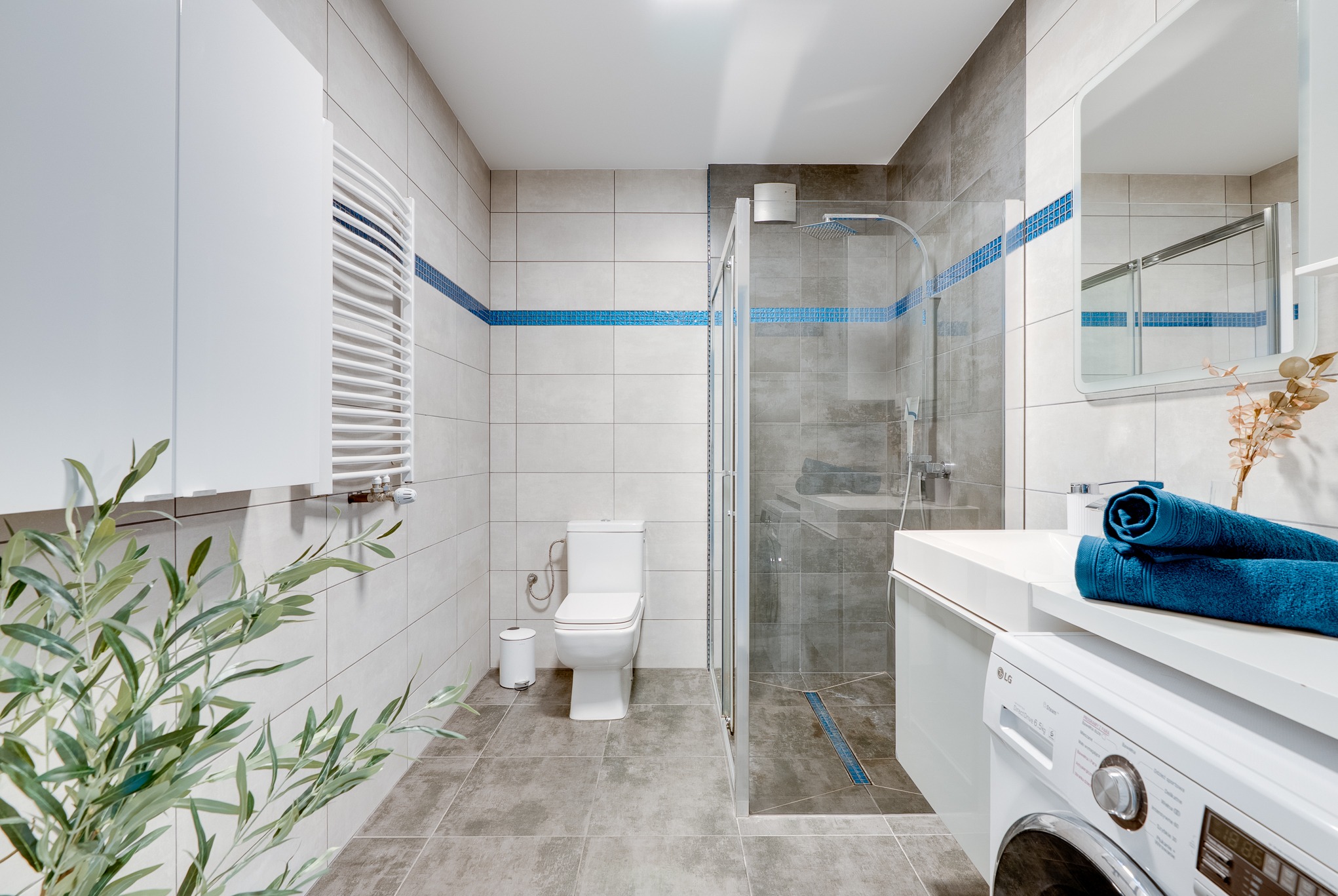 Nowoczesna łazienka z kabiną prysznicową, umywalką z pralką i toaletą. Jasne płytki z niebieskim akcentem. Minimalistyczny design, roślina w rogu.