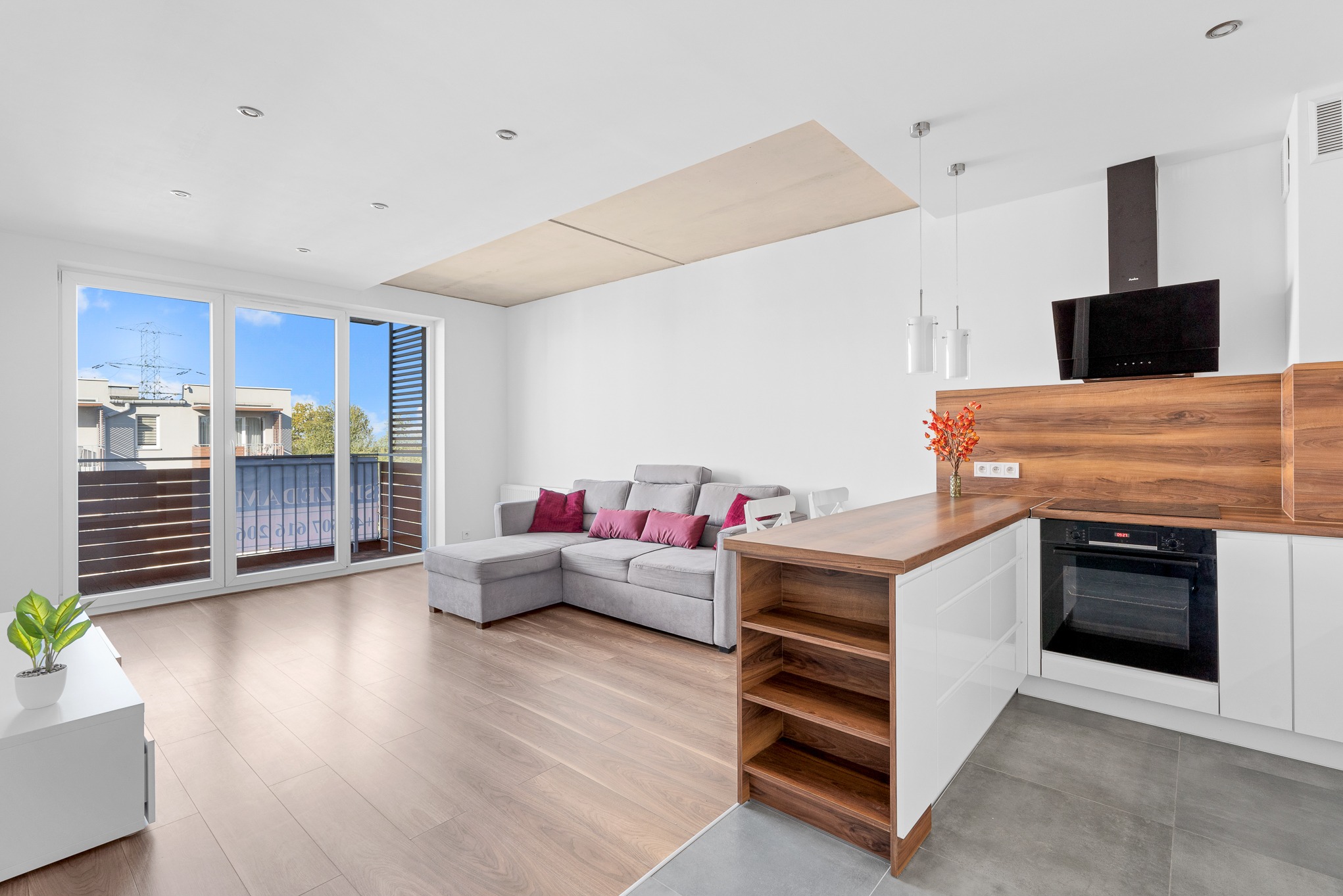 Jasne, nowoczesne wnętrze: salon z szarym narożnikiem i kuchnia z drewnianym blatem. Duże okna balkonowe z widokiem na budynki. Minimalistyczny design.